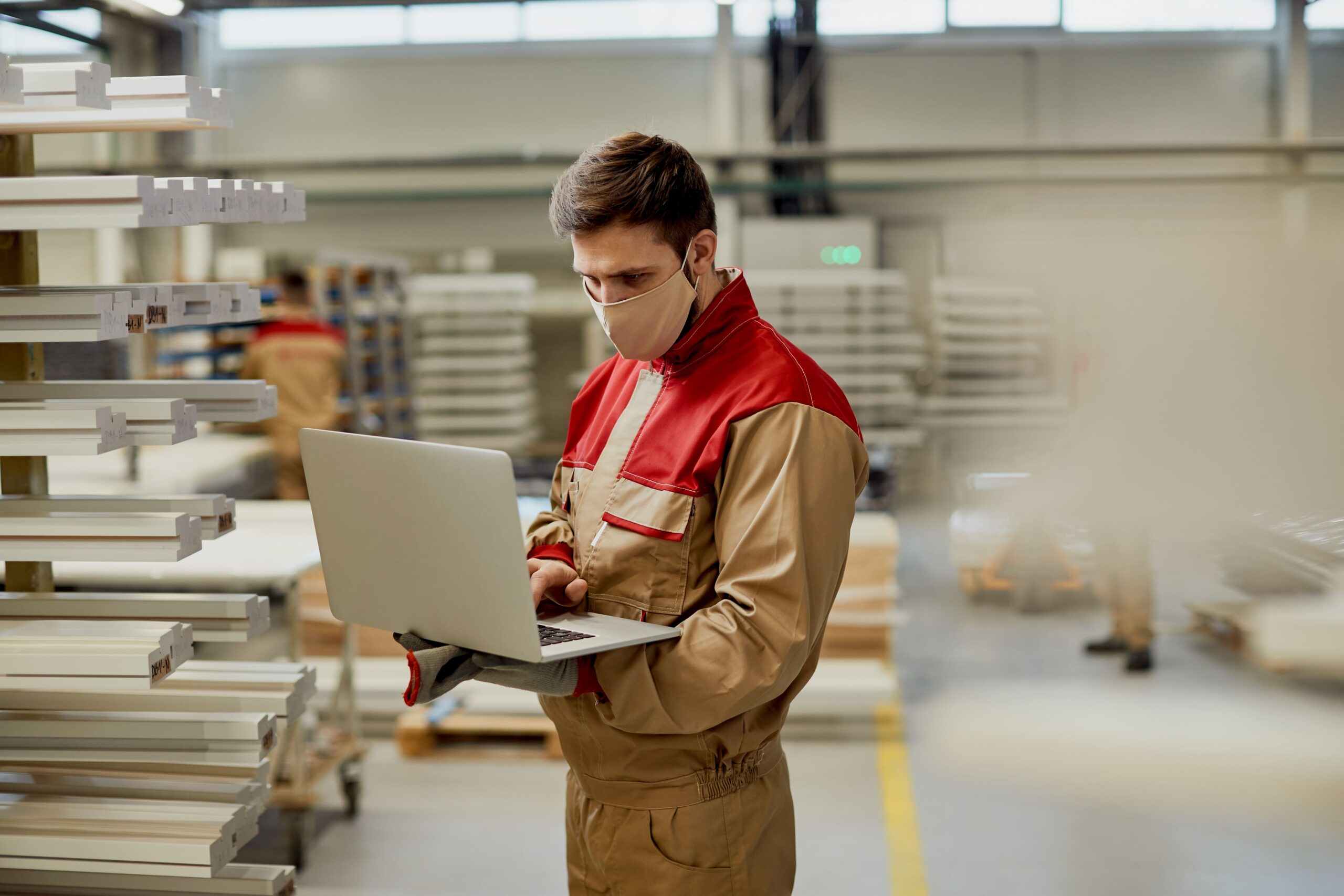 male-carpenter-with-face-mask-using-laptop-while-checking-stock-workshop-min